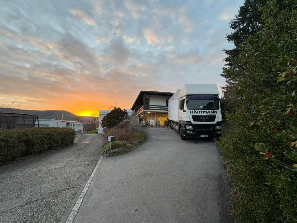 Umzugstransporter in Privateinfahrt in der Schweiz. Im Hintergrund ist ein Sonnenuntergang zu sehen.